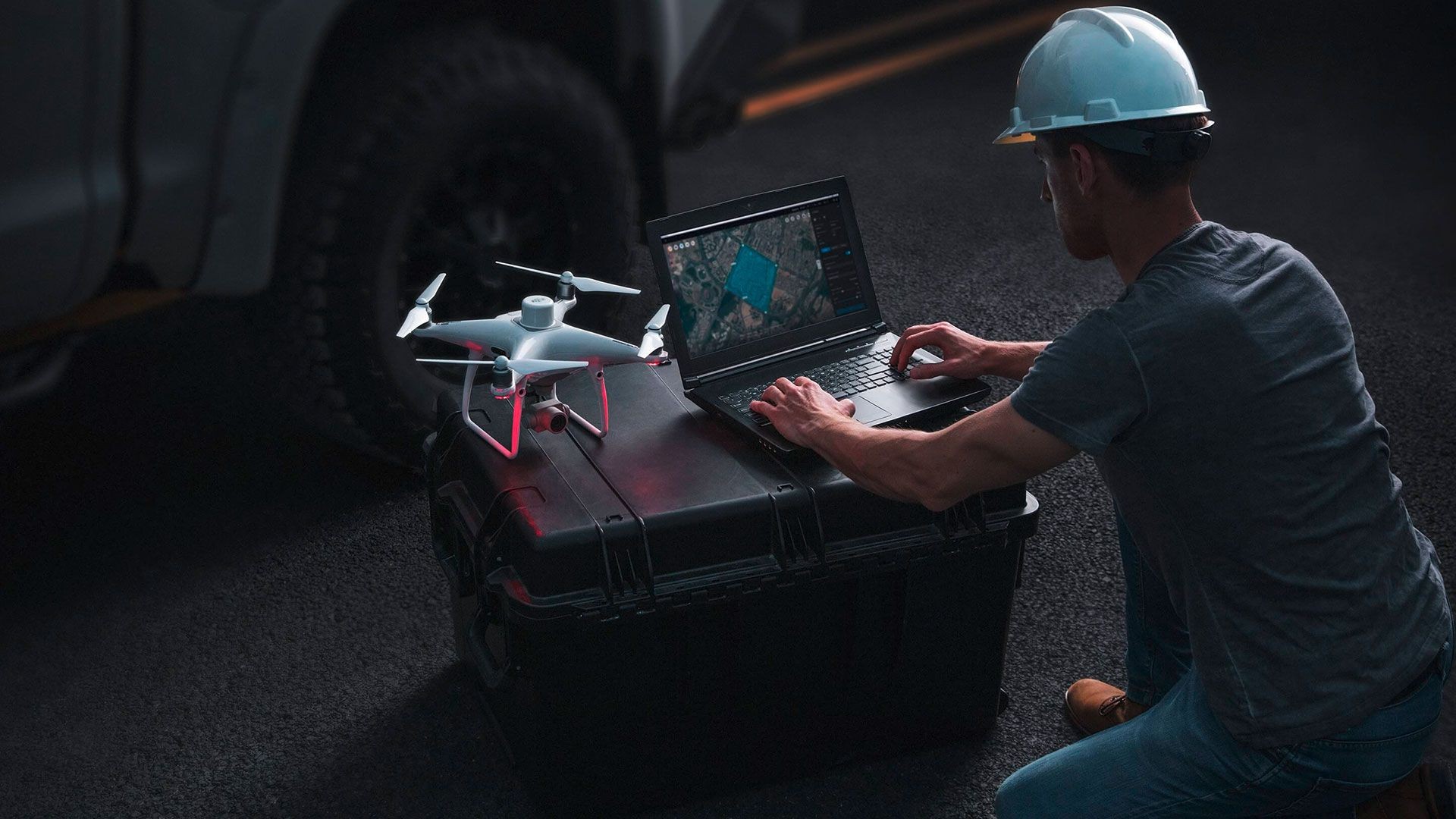 Person in a hard hat operating a laptop next to a drone on a black case, outdoors.