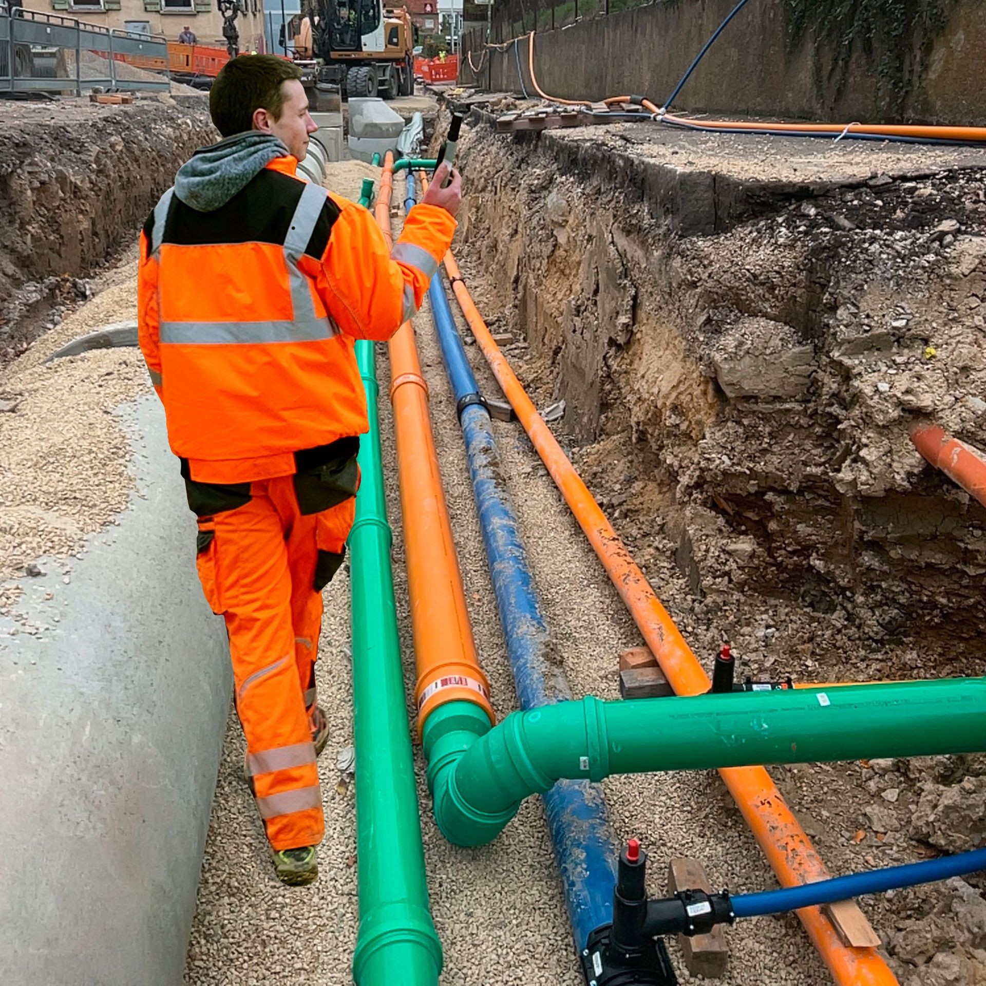 Construction worker in orange gear inspecting underground pipes at a trench site.