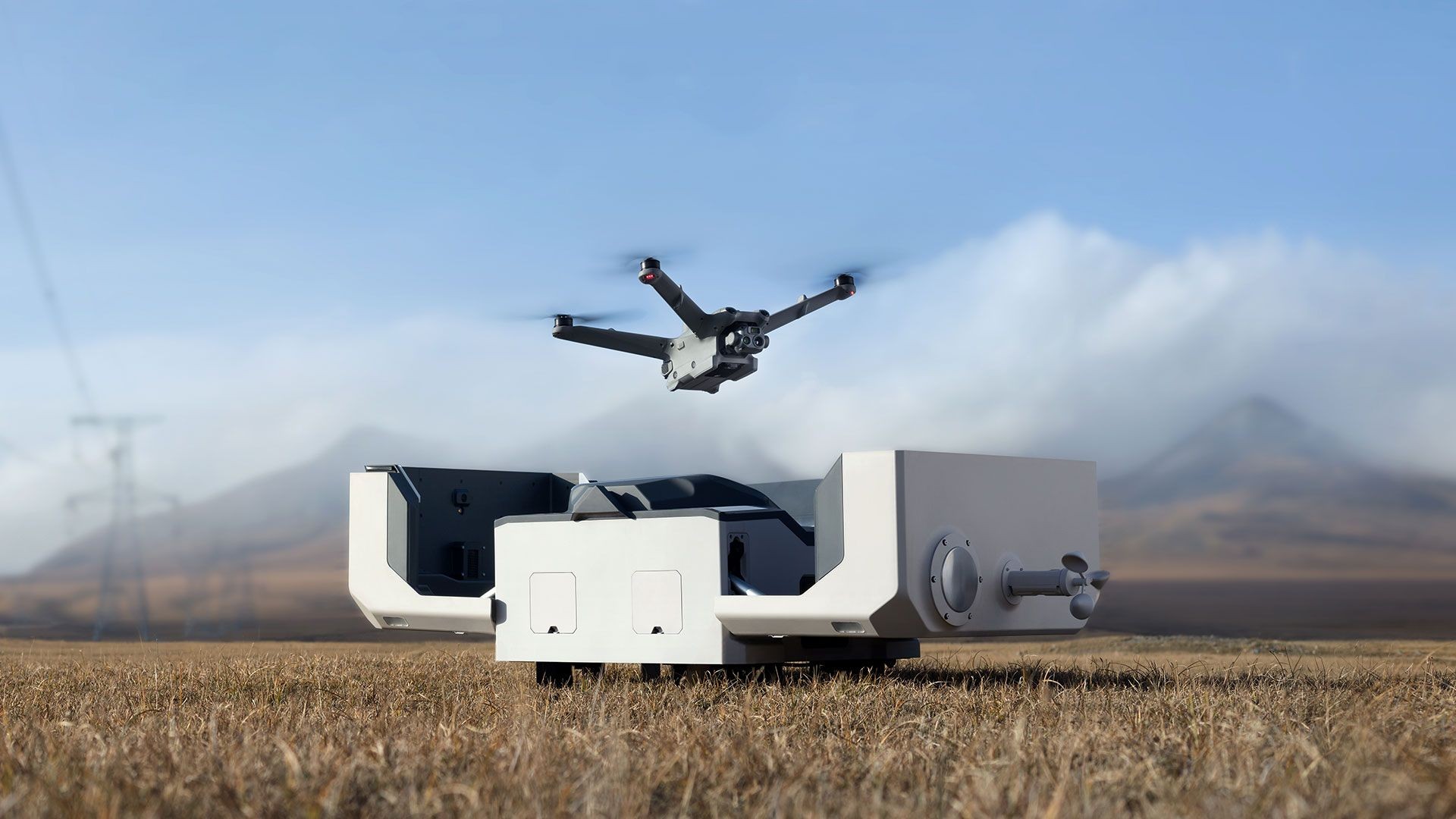 Drone hovering above a docking station in an open field with mountains and power lines in the background.