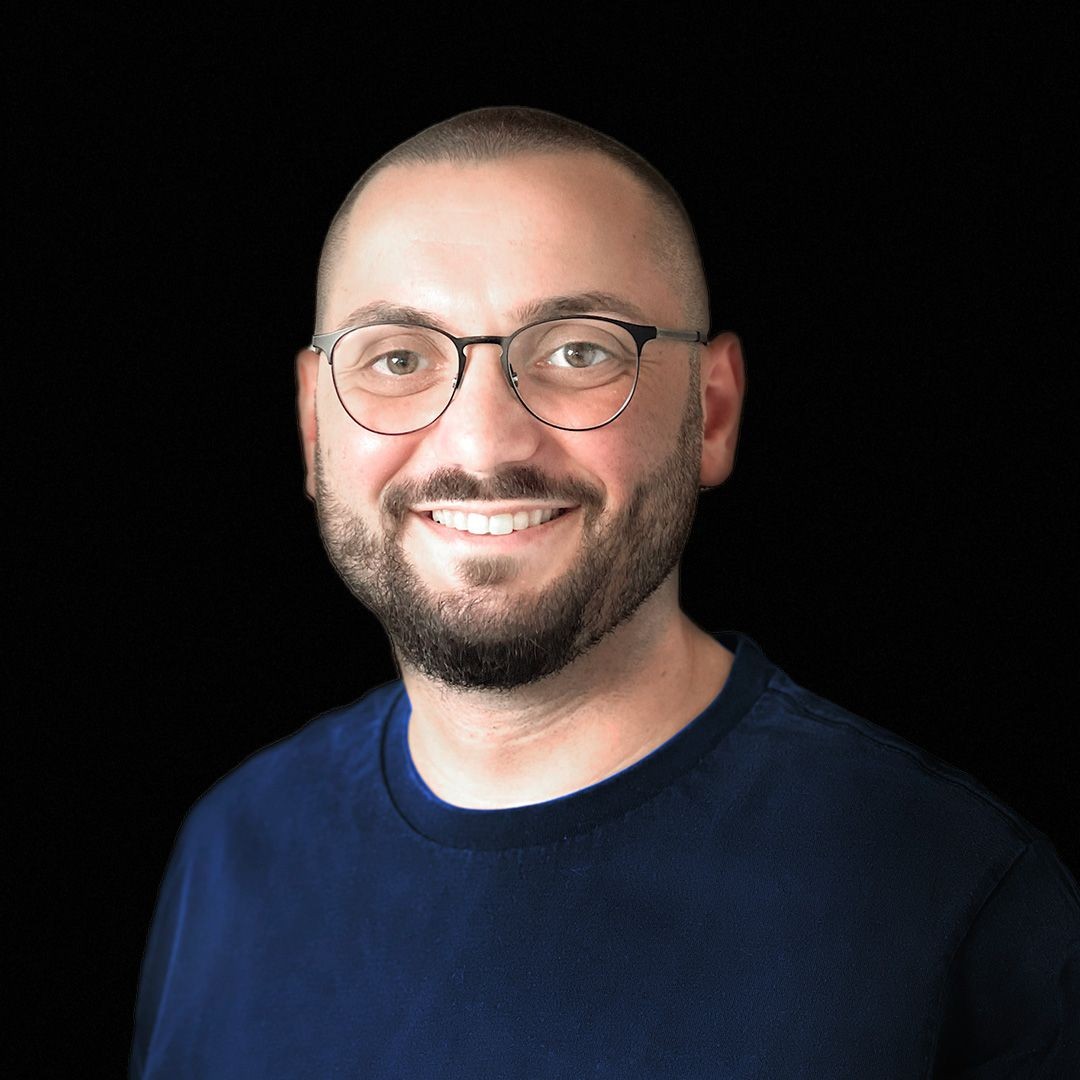 Man with glasses and a beard smiling against a black background, wearing a blue shirt.