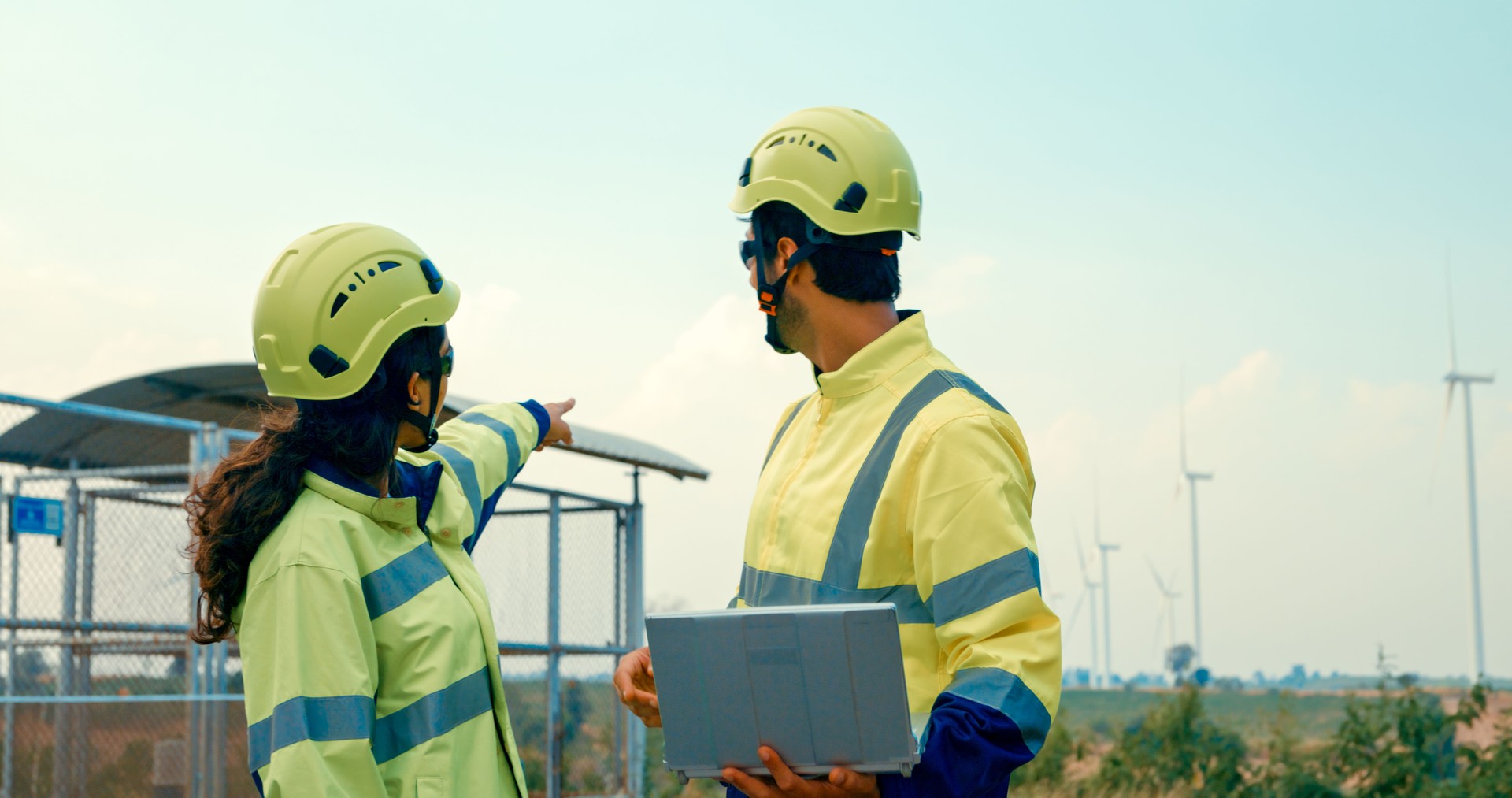 Renewable energy engineers actively engaged in discussion over laptop at power station with wind turbines. Green ecological power generation wind sustainable energy.